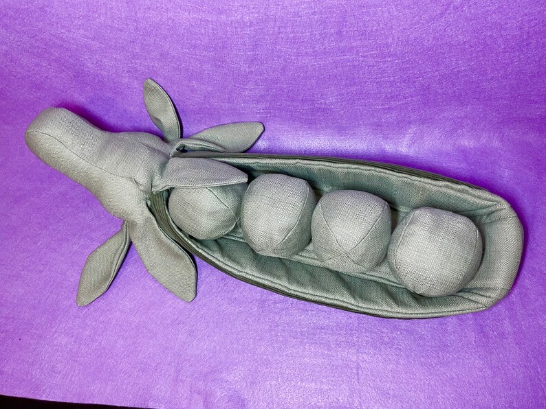 Puede incluir: Una vaina de guisante verde de peluche con cinco guisantes redondos en su interior. La vaina est&aacute; abierta y los guisantes son visibles. La vaina est&aacute; hecha de tela suave y tiene una costura cosida en el centro.