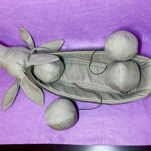 Puede incluir: Una vaina de tela gris con una cabeza de tela gris y cuatro bolas de tela gris. La vaina est&aacute; abierta y las bolas est&aacute;n dentro.