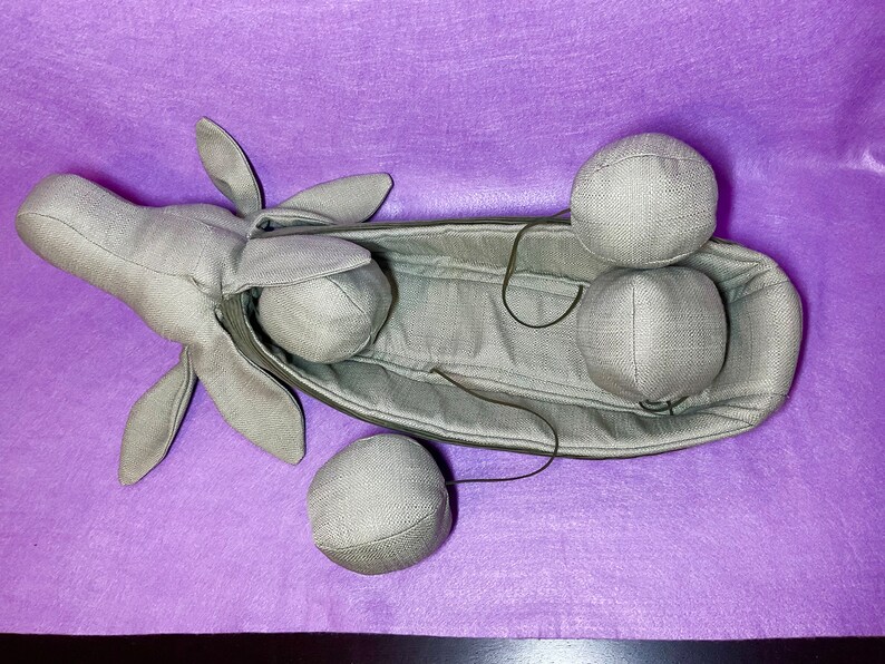 Puede incluir: Una vaina de tela gris con una cabeza de tela gris y cuatro bolas de tela gris. La vaina est&aacute; abierta y las bolas est&aacute;n dentro.