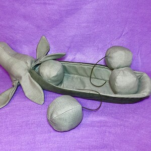 Puede incluir: Una vaina de guisante de tela gris con cuatro guisantes de tela gris en su interior. La vaina de guisante tiene forma de barco con una cabeza y orejas de tela gris en un extremo. Los guisantes est&aacute;n unidos a la vaina con cuerdas negras.