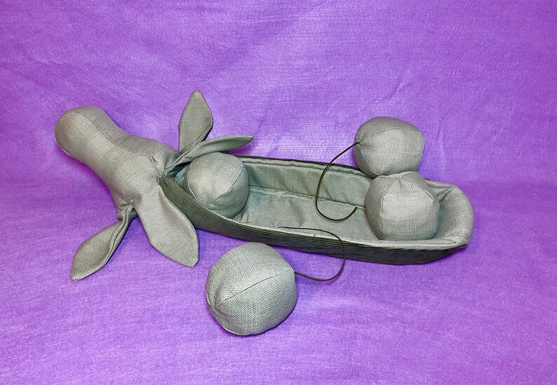 Puede incluir: Una vaina de guisante de tela gris con cuatro guisantes de tela gris en su interior. La vaina de guisante tiene forma de barco con una cabeza y orejas de tela gris en un extremo. Los guisantes est&aacute;n unidos a la vaina con cuerdas negras.