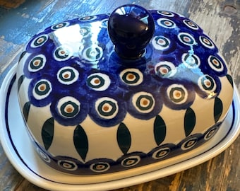 Könnte beinhalten: Eine Keramik-Butterdose mit Deckel, mit einem blau-weißen Blumendesign. Der Deckel hat einen dunkelblauen Knopf. Die Schale steht auf einem weißen Sockel mit blauem Rand. Das Design umfasst blaue Kreise mit braunen und grünen Akzenten.