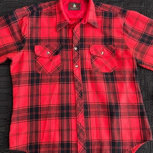 Op de afbeelding: Rood en zwart geruit flanellen overhemd met een knoopsluiting en twee borstzakken. Het shirt heeft een label met de tekst "Heron Bay" met een boomlogo.