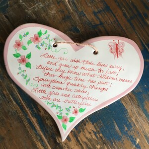 Puede incluir: Una placa de cerámica en forma de corazón rosa con un diseño de mariposa y una cita manuscrita sobre la infancia. La cita dice: "Las niñas pequeñas desean que sus vidas se esfumen, y crecen demasiado rápido; Antes de que sepan lo que significa la infancia, ese tiempo mágico ha pasado; La primavera cambia rápidamente a cielos de verano; Las niñas pequeñas y las orugas pronto se convierten en mariposas."