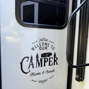 May include: A white camper door with a black decal that reads "Welcome to Our Camper" above the word "Camper" and the names "Kevin & Sarah". The decal includes a camper graphic and two trees.