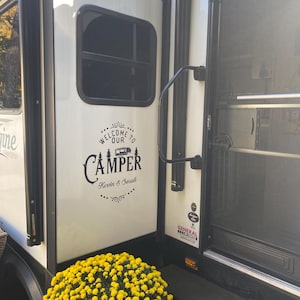 May include: A white camper van with a black decal that says "Welcome to our Camper Kevin & Sarah". The camper van is parked in front of a bush of yellow flowers.
