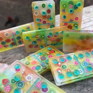 Könnte beinhalten: Ein Set bunter Dominosteine mit einem Frucht-Motiv. Die Dominosteine sind aus klarem Harz gefertigt und haben verschiedene Früchte eingebettet, darunter Erdbeeren, Zitronen, Limetten und Wassermelonen. Die Dominosteine sind in einem zufälligen Muster auf einer Holzoberfläche angeordnet.