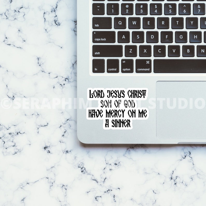 May include: A white sticker with black text on a laptop. The sticker reads "LORD JESUS CHRIST SON OF GOD HAVE MERCY ON ME A SINNER". The laptop is on a white and gray marble surface.