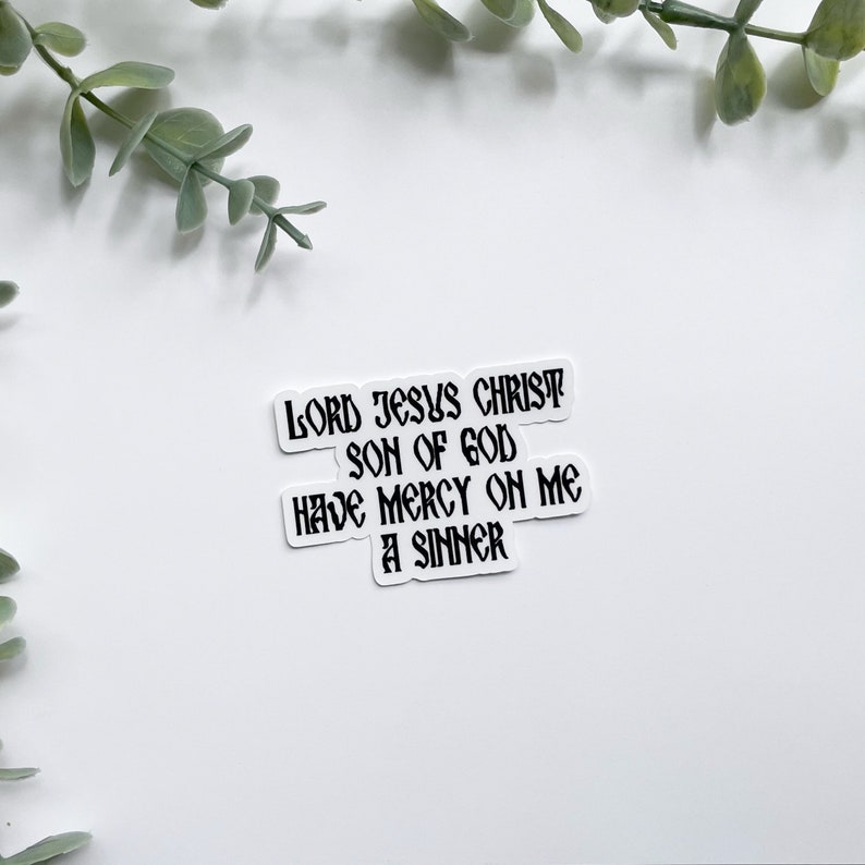 May include: Three white stickers with black text. The top sticker reads "LORD JESUS CHRIST". The middle sticker reads "SON OF GOD". The bottom sticker reads "HAVE MERCY ON ME A SINNER". Greenery is in the background.