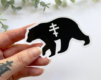 Può includere: Adesivo bianco e nero di un orso con un simbolo di croce sul petto. L'orso cammina verso destra.