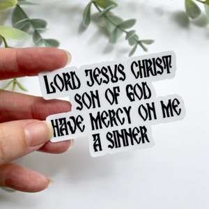 May include: A white sticker with black text that reads "LORD JESUS CHRIST SON OF GOD HAVE MERCY ON ME A SINNER." The sticker is held by a hand against a white background with green foliage.