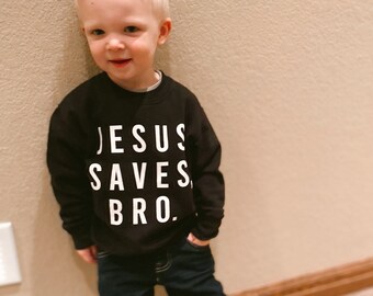 May include: A young boy wearing a black sweatshirt with the text "JESUS SAVES BRO." in white letters. He is also wearing blue jeans and black sneakers.