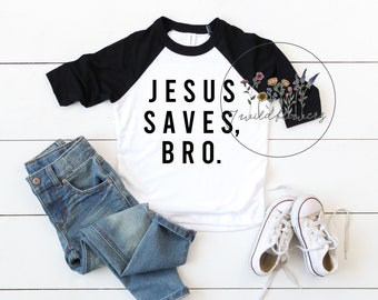 May include: A white and black baseball tee shirt with the text "JESUS SAVES, BRO." printed in black. The shirt is laying flat on a white wood background with a pair of blue jeans and white sneakers.