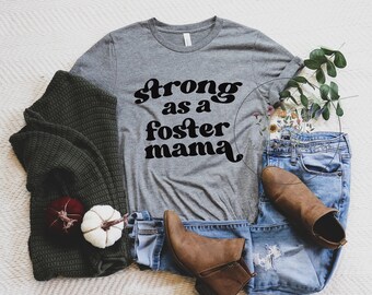 May include: A gray t-shirt with the text "strong as a foster mama" in black retro-style lettering. The shirt is laid out on a white surface with a green sweater, blue jeans, and brown boots.