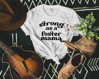 May include: White t-shirt with the text "strong as a foster mama" in black retro font. The shirt is laid out on a green and black patterned surface with a brown hat, a guitar, and brown boots.