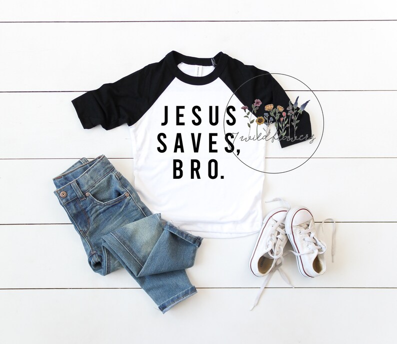May include: A white and black baseball tee shirt with the text "JESUS SAVES, BRO." printed in black. The shirt is laying flat on a white wood background with a pair of blue jeans and white sneakers.