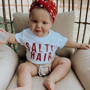 May include: A baby wearing a white t-shirt with the text "Salty Hair" in red lettering. The baby is wearing a red and white polka dot turban and is sitting in a wicker chair.
