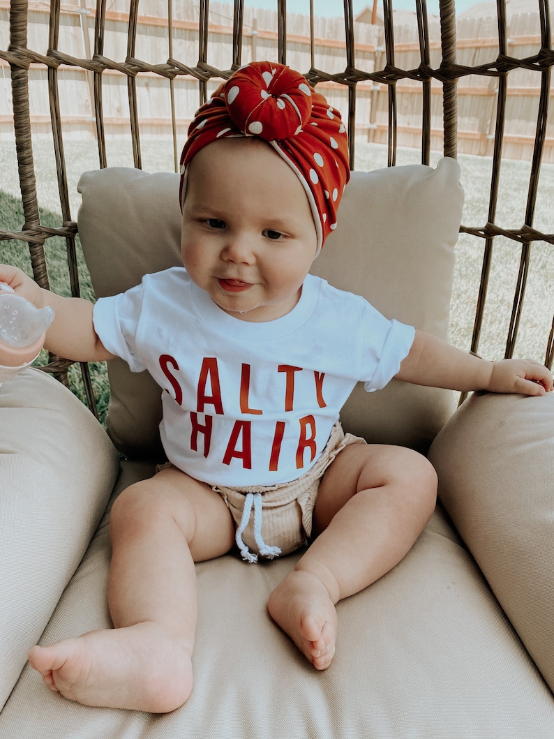 May include: A baby wearing a white t-shirt with the text "Salty Hair" in red lettering. The baby is wearing a red and white polka dot turban and is sitting in a wicker chair.
