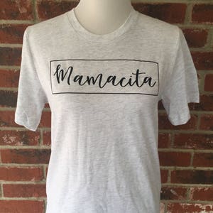 May include: A heather grey t-shirt with a black box and the text "Mamacita" in a cursive font.
