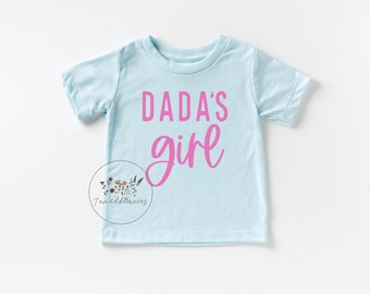 Puede incluir: Una camiseta de manga corta azul claro con el texto "DADA'S girl" en letras rosas.
