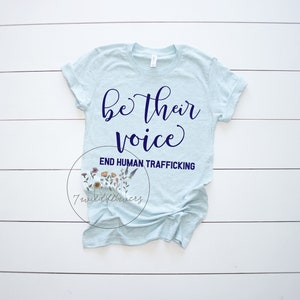 Puede incluir: Una camiseta azul claro con el texto "be their voice" en letras azul marino. Debajo del texto est&aacute; la frase "END HUMAN TRAFFICKING" en letras azul marino. La camiseta est&aacute; colocada sobre una superficie de madera blanca.