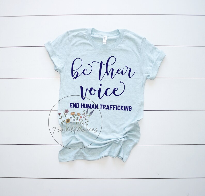 Puede incluir: Una camiseta azul claro con el texto "be their voice" en letras azul marino. Debajo del texto est&aacute; la frase "END HUMAN TRAFFICKING" en letras azul marino. La camiseta est&aacute; colocada sobre una superficie de madera blanca.
