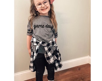 Puede incluir: Una niña pequeña con una camiseta gris con el texto "game day" en letras negras. También lleva pantalones negros, una camisa de cuadros negros y blancos atada a la cintura y zapatillas blancas. Lleva un lazo rosa en el pelo y gafas.