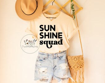 Puede incluir: Una camiseta beige con el texto "SUNSHINE squad" en letras negras. La camiseta está colgada en una percha de madera con un sombrero de paja y un bolso tejido.