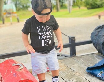 May include: A toddler wearing a black t-shirt with the text "It was Papa's Idea" in white, a black mesh baseball cap, white shorts, and black and white sneakers. The toddler is standing on a wooden surface.