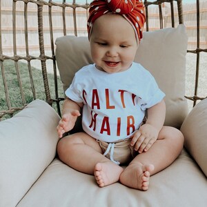 May include: A baby wearing a white shirt with the text "Salt Hair" in red letters, sitting in a wicker chair. The baby is wearing a red and white polka dot turban.