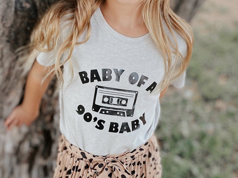 May include: A light gray t-shirt with a black graphic of a cassette tape and the text "Baby of a 90's Baby".