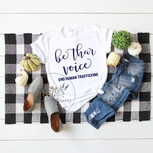 Puede incluir: Camiseta blanca con el texto "be their voice" y "END HUMAN TRAFFICKING" en letras azul marino. La camiseta est&aacute; colocada sobre una superficie a cuadros blancos y negros con un par de zapatos grises, unos vaqueros azules y decoraci&oacute;n de oto&ntilde;o.