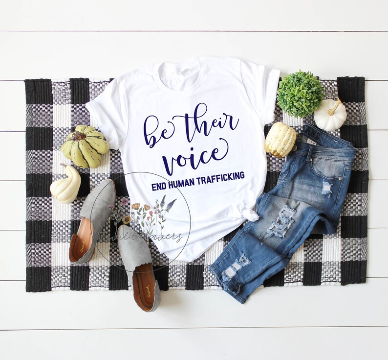 Puede incluir: Camiseta blanca con el texto "be their voice" y "END HUMAN TRAFFICKING" en letras azul marino. La camiseta est&aacute; colocada sobre una superficie a cuadros blancos y negros con un par de zapatos grises, unos vaqueros azules y decoraci&oacute;n de oto&ntilde;o.