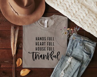 Puede incluir: Una camiseta gris con el texto "Hands Full Heart Full House Full Thankful" impreso en ella. La camiseta está colocada sobre una superficie de madera con un sombrero marrón, un par de vaqueros y algunas espigas de trigo secas.