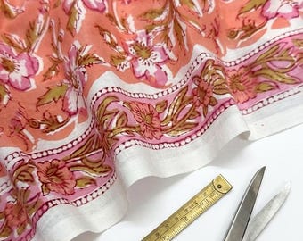 May include: A peach-colored fabric featuring a white and pink floral pattern with green leaves, accompanied by a pair of silver metal scissors and a yellow measuring tape that shows a length of approximately 11 centimeters on the visible scale.