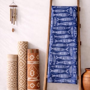 Peut inclure: Un textile bleu avec un motif de poissons blancs est drapé sur une échelle en bois. Des tapis roulés beiges et oranges avec des motifs géométriques sont à proximité. Une cloche à vent est suspendue au mur et un pot en terre cuite est à droite.