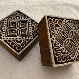 May include: Two square wooden printing blocks with intricate white floral and geometric designs. The blocks are dark brown with white accents, showcasing detailed patterns. These blocks are likely used for textile printing or decorative purposes.