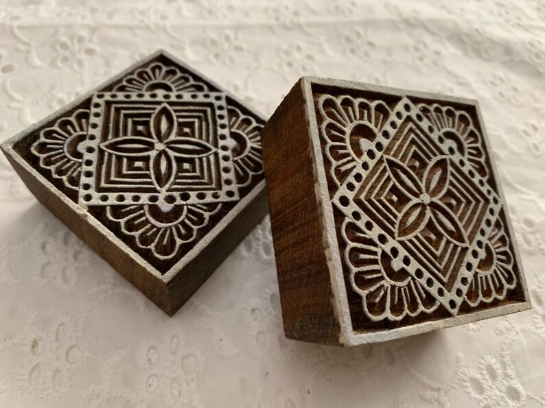 May include: Two square wooden printing blocks with intricate white floral and geometric designs. The blocks are dark brown with white accents, showcasing detailed patterns. These blocks are likely used for textile printing or decorative purposes.