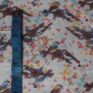 K&ouml;nnte beinhalten: Wei&szlig;er Stoff mit einem sich wiederholenden Muster aus bunten geometrischen Dreiecken und stilisierten V&ouml;geln. Die V&ouml;gel sind in Blaut&ouml;nen, Gelb und Braun gehalten und sitzen auf &Auml;sten mit rosa Bl&uuml;ten. Ein blaues Ma&szlig;band ist auf der linken Seite des Bildes sichtbar.