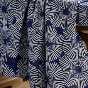 May include: A fabric tablecloth featuring a repeating pattern of large, white, radiating circular floral motifs set against a deep navy blue background. The textile drapes over the edge of a surface, displaying folds and a soft, matte texture.