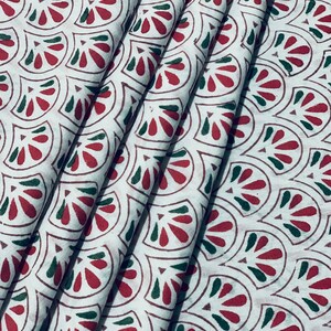 May include: White fabric with a repeating pattern of red and green fan-shaped designs. The fabric is folded, showing the pattern's repeating nature. The design is printed on the fabric.