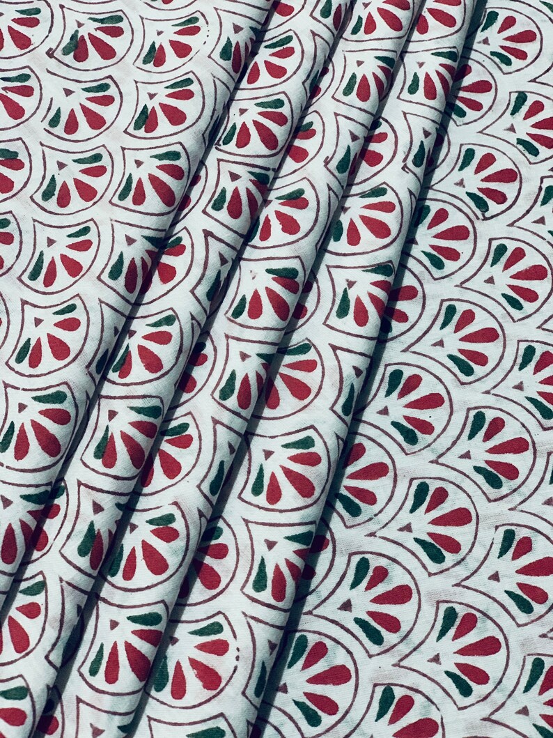 May include: White fabric with a repeating pattern of red and green fan-shaped designs. The fabric is folded, showing the pattern's repeating nature. The design is printed on the fabric.