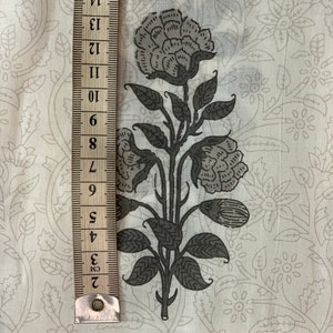 May include: White fabric with a gray floral print. The print features a large flower with multiple petals and leaves. A ruler is visible on the left side of the image.