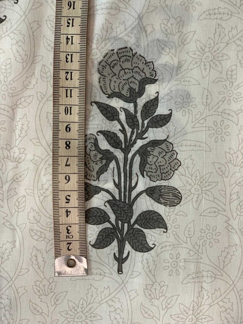 May include: White fabric with a gray floral print. The print features a large flower with multiple petals and leaves. A ruler is visible on the left side of the image.