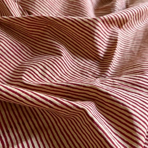 May include: Close-up of a fabric with a repeating pattern of burgundy stripes on a cream background. The fabric is folded and draped, creating shadows and texture. The stripes are evenly spaced and run diagonally across the image.