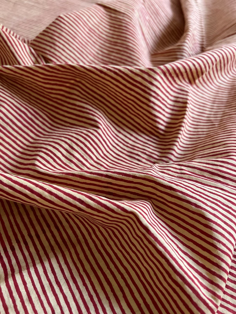 May include: Close-up of a fabric with a repeating pattern of burgundy stripes on a cream background. The fabric is folded and draped, creating shadows and texture. The stripes are evenly spaced and run diagonally across the image.