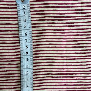 May include: A measuring tape with centimeter markings is placed vertically against a fabric with a cream background and burgundy horizontal stripes. The tape shows measurements up to 20 cm, or approximately 7.87 inches.