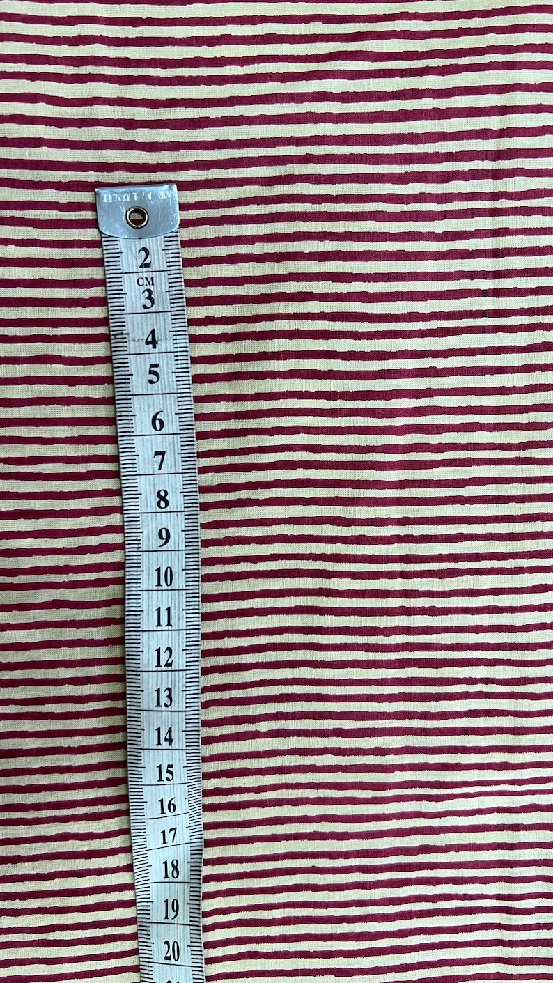 May include: A measuring tape with centimeter markings is placed vertically against a fabric with a cream background and burgundy horizontal stripes. The tape shows measurements up to 20 cm, or approximately 7.87 inches.