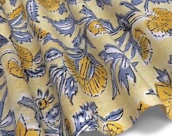 May include: A piece of fabric featuring a repeating floral pattern with yellow and blue flowers on a pale yellow background. The textile has a soft, draped appearance with visible folds that create depth and texture across the surface.