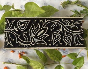 May include: A black and white wooden printing block with a floral design featuring a large flower, leaves, and hearts. The block is on a white fabric background.